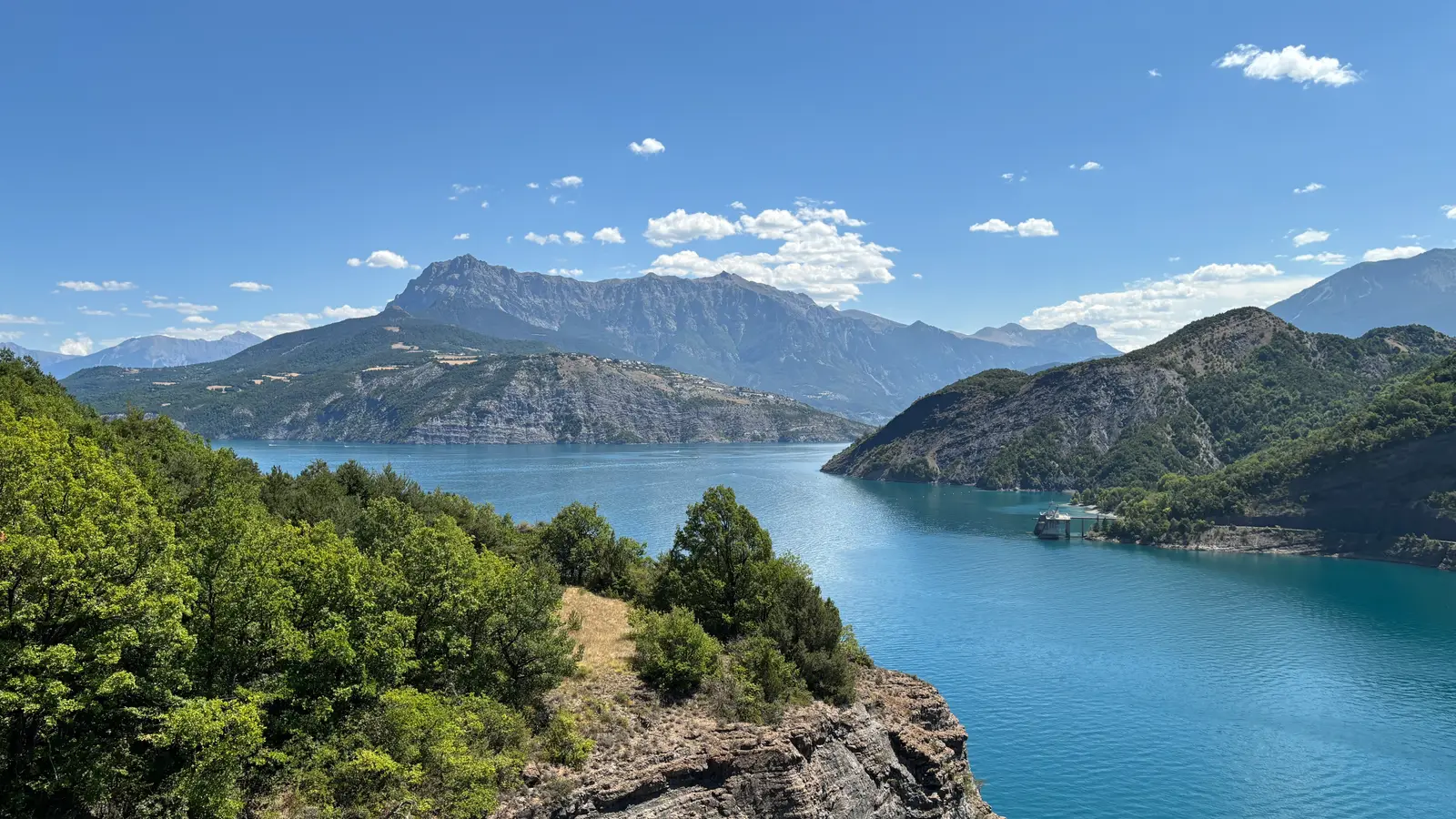 Un grand lac entre des collines boisées, des montagnes au loin, en camaïeu de bleu.